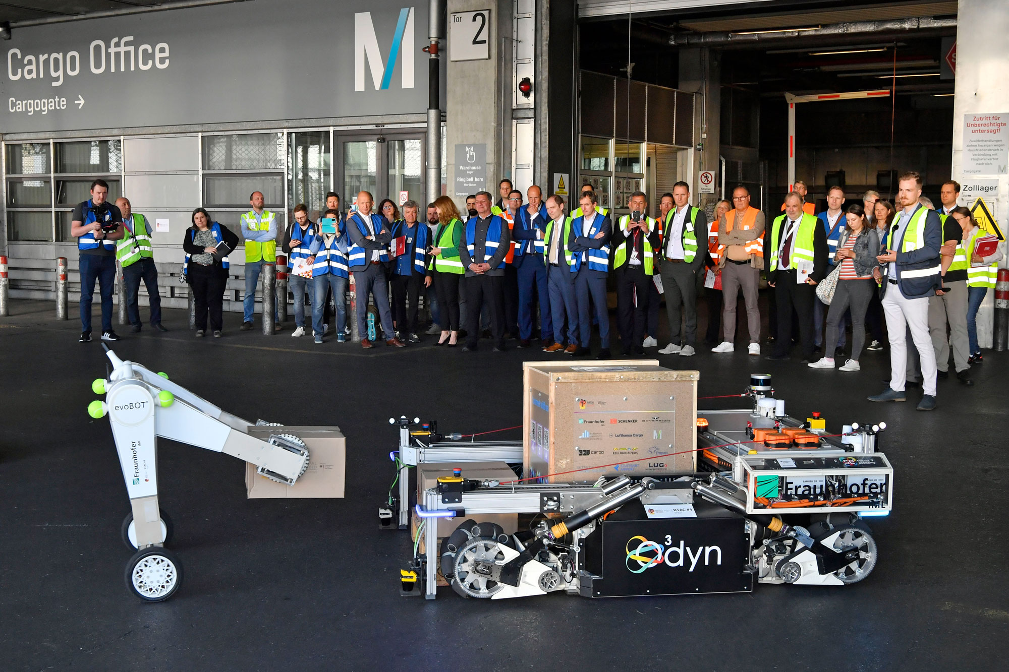 Roboter, einschließlich des Roboterhunds Spot, des omnidirektionalen Transportroboters O³dyn und des evoBots, bei der Luftfrachtabfertigung am Flughafen München, umgeben von Branchenakteuren im Hintergrund während der Präsentation des Forschungsprojekts DTAC.